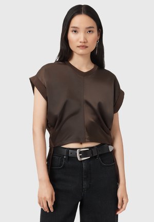 Giovane donna con lunghi capelli scuri che indossa un top corto senza maniche marrone, una cintura nera e jeans neri, in piedi contro uno sfondo semplice.