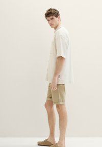 Cămașă scurtă de culoare bej deschis cu un buzunar pe piept, purtată împreună cu pantaloni scurți de culoare bej și pantofi slip-on maro, purtată de un model tânăr de sex masculin.