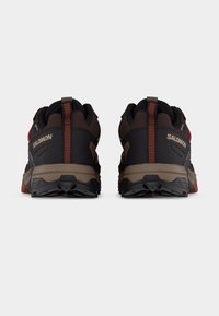 Ζευγάρι μαύρων και καφέ παπουτσιών πεζοπορίας Salomon με κόκκινες λεπτομέρειες, εμφανιζόμενα από πίσω σε λευκό φόντο.