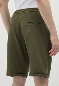 Uomo che indossa pantaloni corti verde oliva fino al ginocchio con orli arrotolati e cintura elastica, mostrato da dietro.