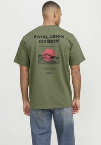 Camiseta de algodón verde oliva de mangas cortas. El diseño gráfico presenta un sol rosa y montañas. El texto dice "Royal Denim Division" por encima del gráfico.
