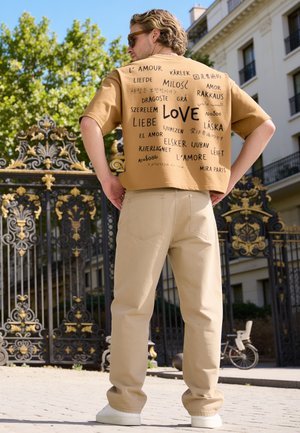 Uomo con occhiali da sole e pantaloni beige davanti a un cancello nero e oro ornato, indossa una camicia color sabbia con "love" scritto in più lingue.