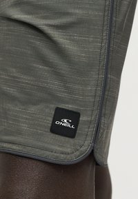 Κοντινή λήψη γκρι σορτς με σχέδια και μαύρο πατς λογότυπου O'Neill σε σκουρόχρωμο πόδι, εμφανίζονται η πλαϊνή ραφή και οι λεπτομέρειες του τελειώματος.