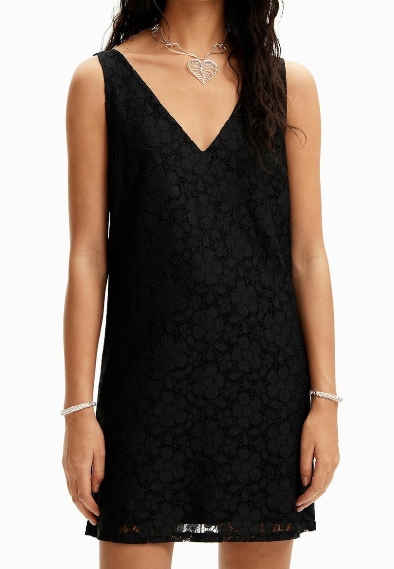 Robe en dentelle noire avec décolleté en V, design sans manches, motif floral et ourlet à hauteur du genou. Portée avec un collier et un bracelet en cœur argenté.