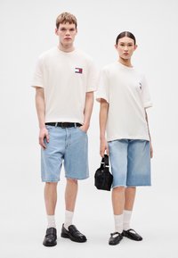 Duas pessoas vestem roupas casuais: t-shirts off-white, calções de ganga azul claro, sapatos pretos e uma pequena bolsa preta. Design e cores simples.