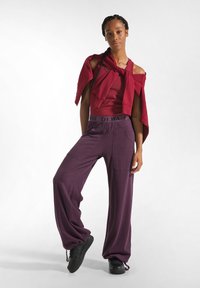 Rode off-shoulder top gelaagd met een bijpassend rood kledingstuk, gecombineerd met loszittende paarse broek met zakken en zwarte sneakers.