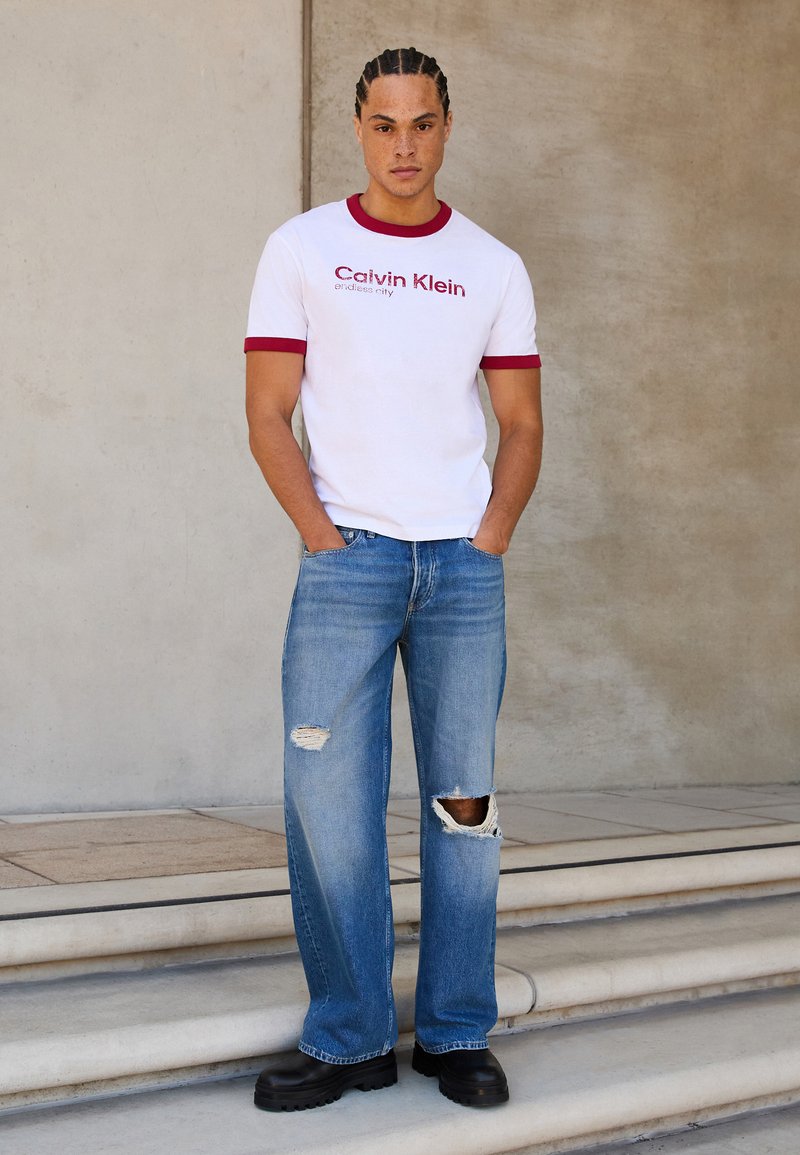 Bijela majica s crvenim detaljima i "Calvin Klein" logom, u kombinaciji s poderanim plavim trapericama i crnim čvrstim cipelama, stoji na stepenicama.