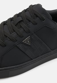 Sneaker nera con un pannello laterale testurizzato, lacci neri piatti e un badge a forma di triangolo. Il materiale appare liscio con una finitura opaca.