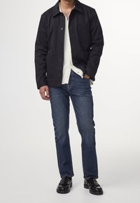 Hombre vestido con chaqueta oscura de color marino, camisa blanca abotonada, jeans azules y mocasines de cuero negros, de pie con una mano en el bolsillo.