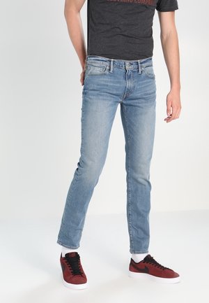 Personne portant un jean slim bleu clair, des baskets noires et rouges avec des semelles blanches, et un t-shirt gris foncé sur un fond uni.