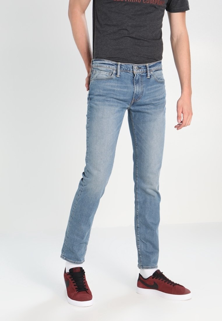 Personne portant un jean slim bleu clair, des baskets noires et rouges avec des semelles blanches, et un t-shirt gris foncé sur un fond uni.