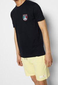 Námořnická modrá bavlněná trička s krátkým rukávem, s kulatou vyšívanou nášivkou v červené, zelené a bílé barvě na hrudi.