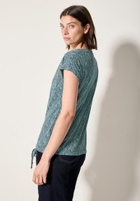 Haut bleu-vert à manches courtes avec un motif de feuilles, encolure ronde et détail de nœud sur le côté. Tissu doux avec une coupe décontractée.