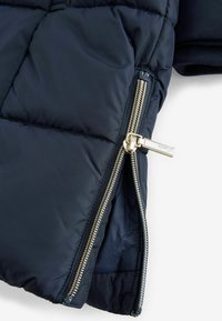 Tamno plavi puffer kaput s zlatnim patentnim zatvaračima, quiltanom teksturom, bočnim džepovima i rebrastim manžetama. Glatka tkanina s sjajnim završetkom.