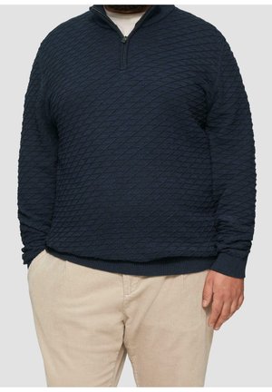 Homme portant un pull texturé bleu marine à fermeture éclair au quart et un pantalon en velours beige, debout avec une main dans la poche devant un fond uni.
