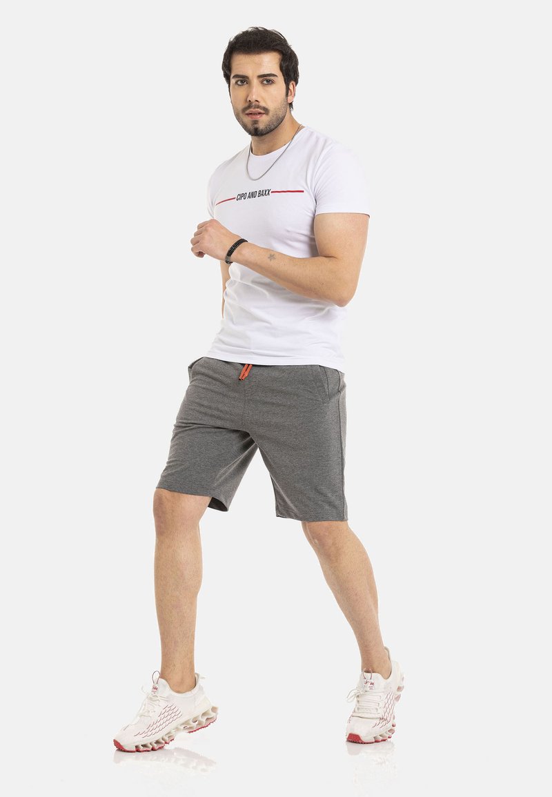 Weißes T-Shirt mit kurzen Ärmeln, darauf schwarzer und roter Text, kombiniert mit grauen Shorts mit seitlichen Taschen und einem orangefarbenen Kordelzug, getragen mit weißen Turnschuhen.
