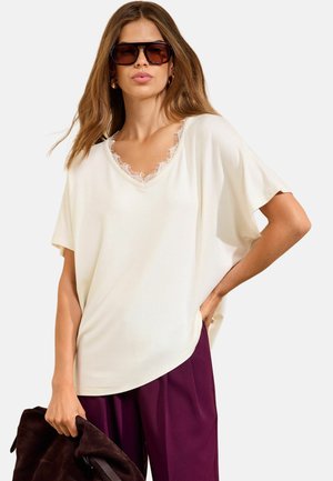 Kvinde iført oversized hvid satin top med blonde detaljer, lilla bukser, store mørke solbriller, holder en mørk tekstureret taske mod hvid baggrund.