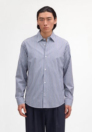 Giovane uomo con camicia a righe verticali blu e bianche e pantaloni scuri, in piedi davanti a uno sfondo bianco uniforme.