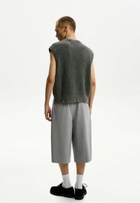 Maglione grigio senza maniche in maglia con motivo testurizzato, abbinato a pantaloni corti grigi chiari fino al ginocchio e scarpe nere, visto da dietro.