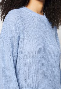 Ljust blå stickad tröja med lös passform, rund halsringning och ribbade ärmslut. Texturen är mjuk och något genomskinlig.