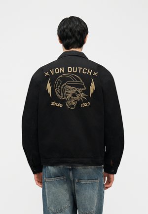 Oseba v črni jakni z bež vezeno pantero s čelado in napisom "VON DUTCH since 1929" na hrbtni strani.