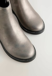 Metalen zilveren enkellaarsjes met een gladde afwerking, ronde neus en zwarte zool met decoratieve metallic accenten langs de rand.