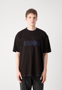 Melna, pārāk liela T-kreklu ar zilu izšuvumu un izteiksmīgu "HUGO" logotipu priekšā. Apaļš dekoltē un īsas piedurknes.