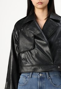 Veste en cuir noire cropped avec un motif texturé en relief, un large col et des détails de boutons, associée à un jean bleu taille haute.