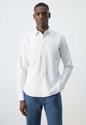 Hombre de pie vestido con una camisa blanca de manga larga abotonada y vaqueros azules contra un fondo claro y liso, con una mano en el bolsillo.
