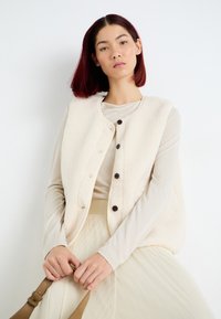 Kremfarget fleecevest med fem mørke knapper, kombinert med en lys tan topp og et plissert, lys beige skjørt. Glatt tekstur, rund hals.