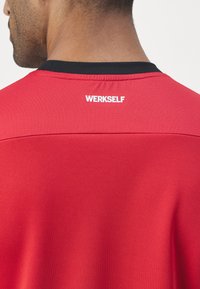 Camisola atlética vermelha com um colarinho redondo preto, apresentando um tecido texturizado e o logótipo "WERKSELF" em branco na parte de trás do colarinho.