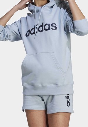 Persoon in een lichtgrijze Adidas hoodie en bijpassende shorts met zwarte Adidas branding aan de voorkant en zijkant.