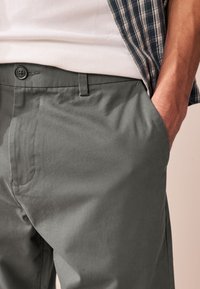 Homme portant un pantalon gris avec la main dans la poche, un t-shirt blanc côtelé et une chemise à carreaux partiellement visible sur fond neutre.