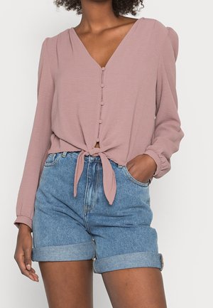 Kvinde iført lavendelfarvet bluse med lange ærmer og knapper foran, bundet ved taljen, og lyseblå denimshorts med manchetter, hånd i lommen.