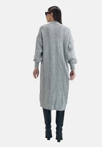 Grå stickad cardigan med vertikal ribbstruktur, översized ärmar och hög krage, som parats med åtsittande svarta stövlar. Enhetlig textur genomgående.