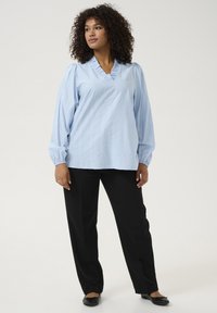Blouse bleu clair à manches longues avec un col en V froncé et un tissu texturé, associée à un pantalon noir à jambe droite et des chaussures plates noires.