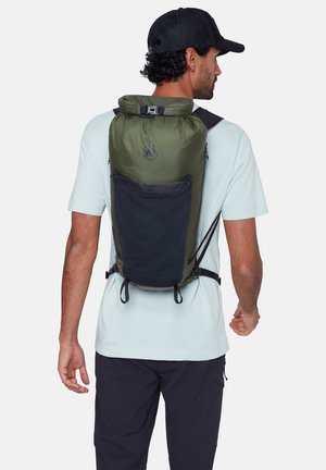 Homme portant une casquette noire, une chemise bleu clair et un pantalon noir, portant un sac à dos à enroulement vert et noir avec poche avant, vu de dos.