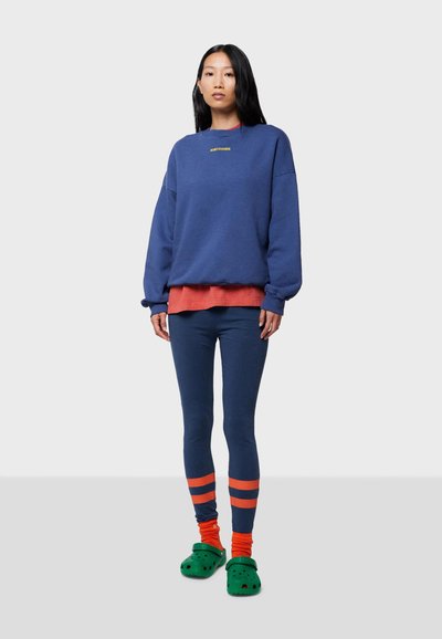Blå sweatshirt med gul text, lager över en korallfärgad skjorta. Blå leggings, strumpor med orange-linjer och gröna träskor. Enkelt, avslappnat ensemble.