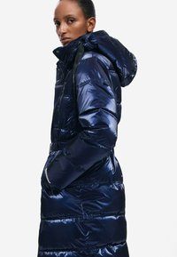 Navyblauwe gewatteerde jas met een glanzend, gewatteerd ontwerp, oversized capuchon en ritssluiting. Voorzien van zijzakken en een ceintuur om de taille.