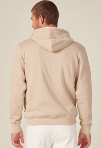 Felpa beige con una finitura testurizzata, cappuccio con cordino e polsini a coste. Cuciture visibili e vestibilità rilassata. Branding minimal sul lato.