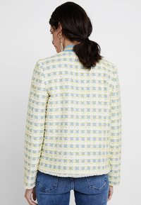 Texturierter Tweed-Jacke in Pastellgelb und Blau mit quadratischem Muster, rundem Ausschnitt und Fransenverzierungen an den Kanten.