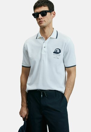 Witte polo met marineblauwe accenten op de kraag en mouwen. Voorzien van een geborduurd logo en een knopenlijst. Donkere broek en een zonnebril gedragen.