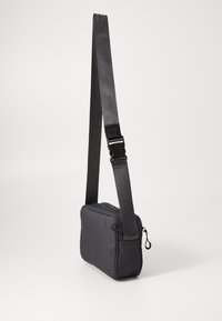 Schwarze Stoff-Umhängetasche mit rechteckiger Form, Reißverschluss und verstellbarem Trageriemen mit metallischem Schnallenverschluss. Strukturierte Oberfläche.
