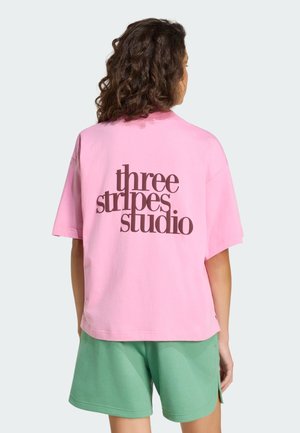 Różowy bawełniany t-shirt o luźnym kroju z brązowym napisem graficznym „three stripes studio” na plecach. Noszony z zielonymi szortami.