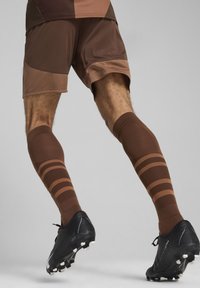 Shorts de sport marron associés à des chaussettes marron jusqu'aux genoux avec des rayures horizontales plus foncées, et des chaussures à crampons noires avec des tiges blanches.