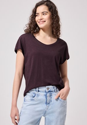 Jeune femme aux cheveux bouclés porte un t-shirt violet foncé à manches courtes et un jean bleu clair, souriant en regardant de côté, main dans la poche.