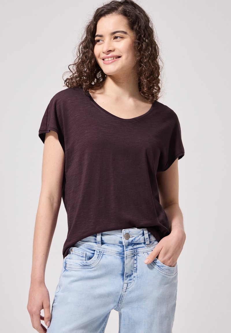 Jeune femme aux cheveux bouclés porte un t-shirt violet foncé à manches courtes et un jean bleu clair, souriant en regardant de côté, main dans la poche.