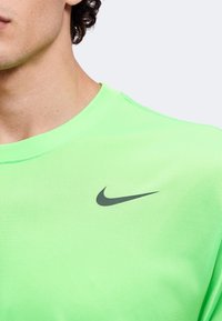Άνδρας που φορά φωτεινό πράσινο μπλουζάκι Nike με μαύρο λογότυπο Nike swoosh στο αριστερό στήθος, εμφανιζόμενος από τον λαιμό έως τη μέση του κορμού.