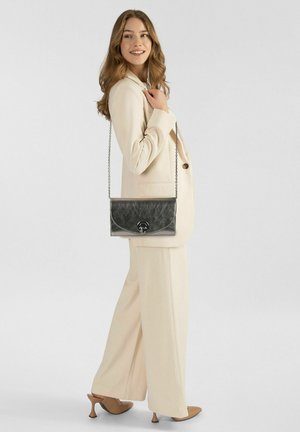 Borsa a mano in pelle grigia con tracolla a catena, dotata di chiusura con patta arrotondata e logo in rilievo, indossata con un completo beige chiaro.