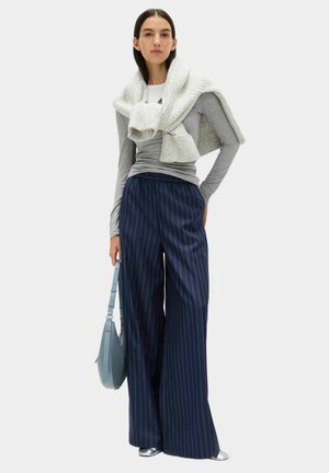 Donna con maglia a maniche lunghe grigia e maglione crema a maglia drappeggiato sulle spalle, pantaloni blu navy a righe larghe, scarpe argento e borsa blu in mano.